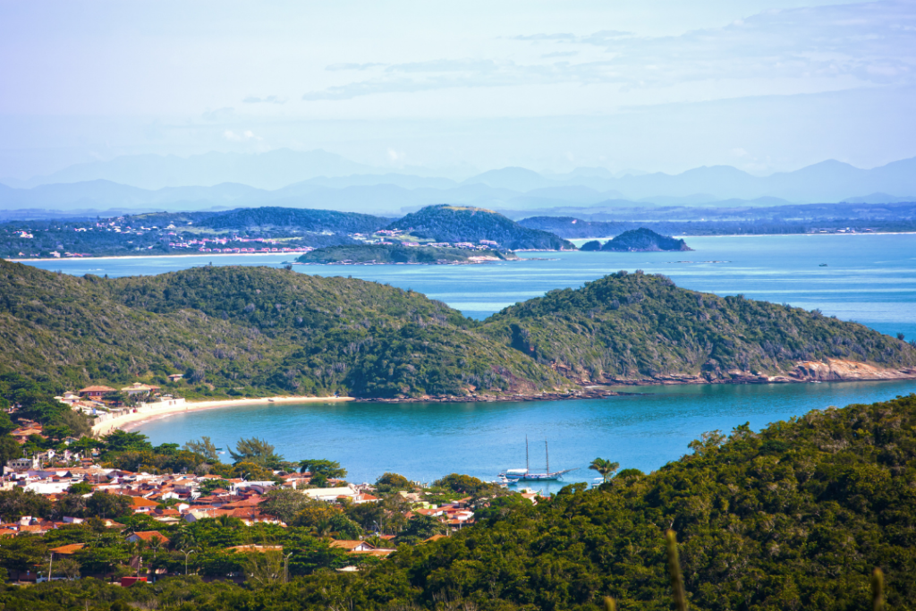 Vista panorâmica da cidade de Búzios, um dos destinos mais promissores do mundo. É possível ver morros com vegetação, mar e algumas casas.