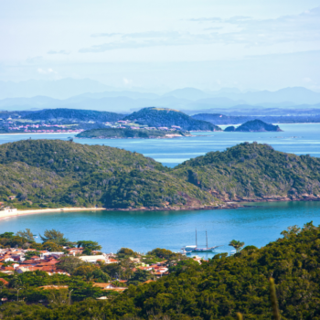 Vista panorâmica da cidade de Búzios, um dos destinos mais promissores do mundo. É possível ver morros com vegetação, mar e algumas casas.