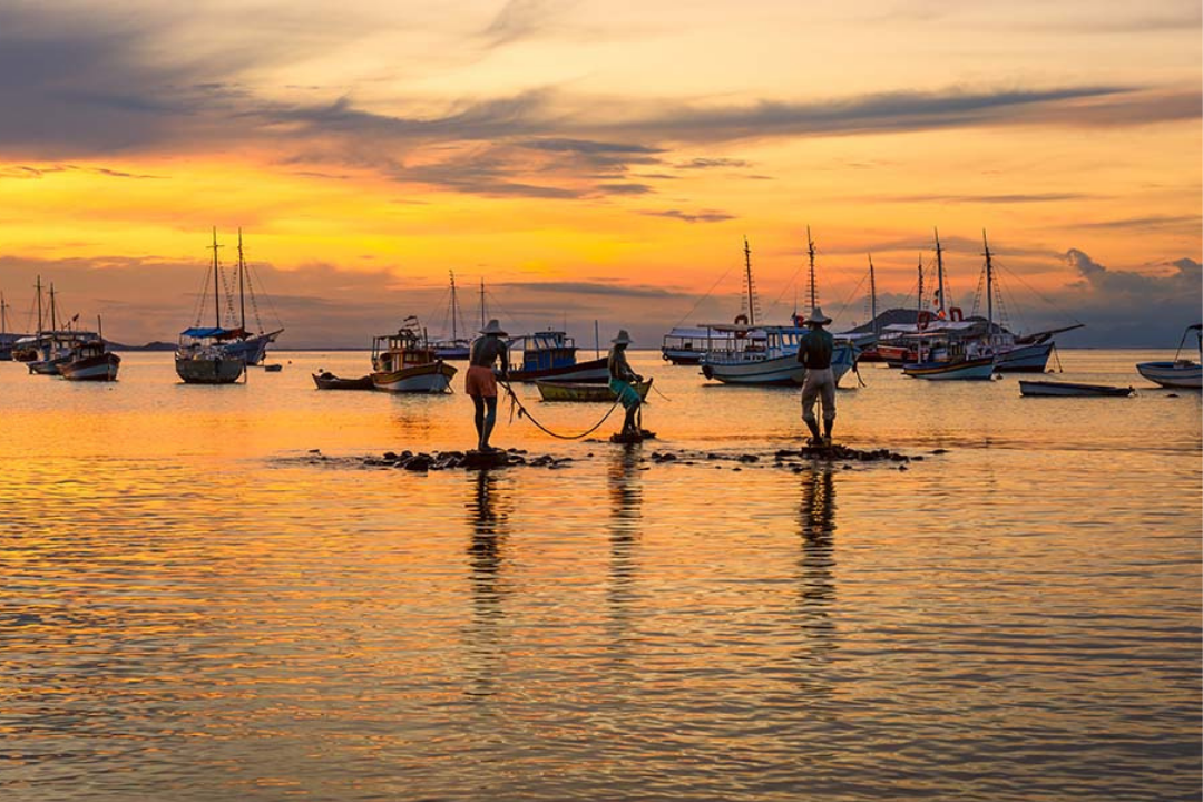 Monumento dos Três Pescadores, um dos símbolos da cidade de Búzios. Final de tarde com pôr do sol, diversas embarcações atrás da escultura.