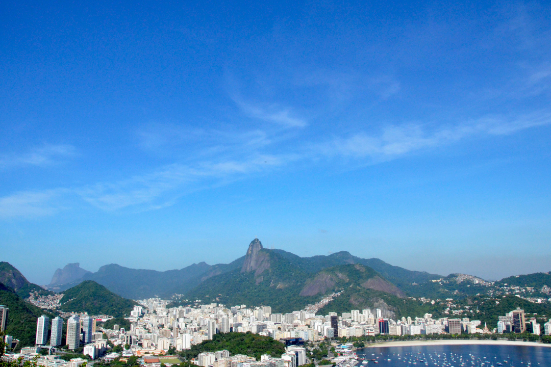 Vista do topo do Morro da Urca, no alto do seus 220 metros de altura. É possível ver o céu azul, parte da Baía de Guanabara, muitos prédios e a vegetação que fazem parte do Rio de Janeiro.