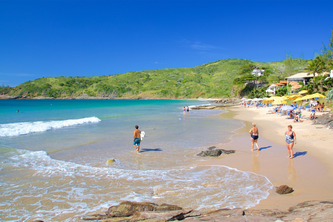 Praia Brava, em um dia de sol, com algumas pessoas na faixa de areia e outra adentrando o mar com uma prancha. Guarda-sóis e um morro com vegetação ao fundo.