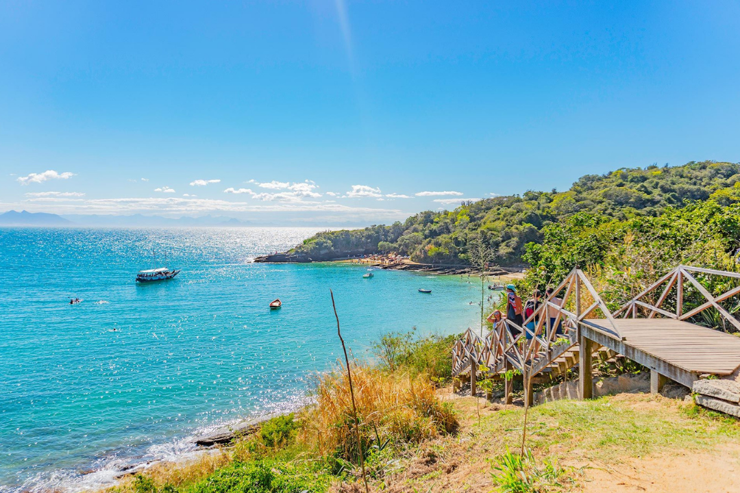 Vista da entrada da Praia da Azeda, que contém uma escadaria para chegar até o local. É possível ver o mar cristalino com uma embarcação e algumas canoas, além de toda a vegetação ao redor do mar.