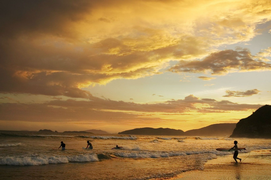 Praia de Geribá, uma das mais famosas da cidade de Búzios. Pôr do sol, sinalizando final de tarde, com alguns banhistas no mar e um surfista caminhando em direção a ele com sua prancha.