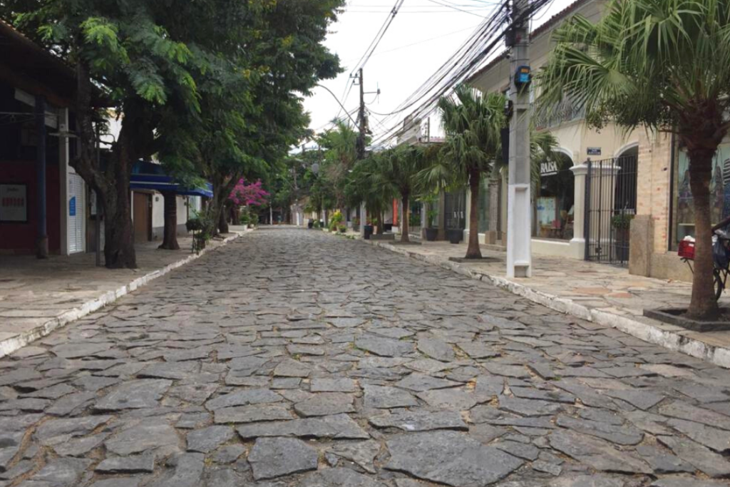 Imagem da Rua das Pedras vazia, sem pessoas na foto. Local com muitas árvores nas calçadas de ambos os lados.