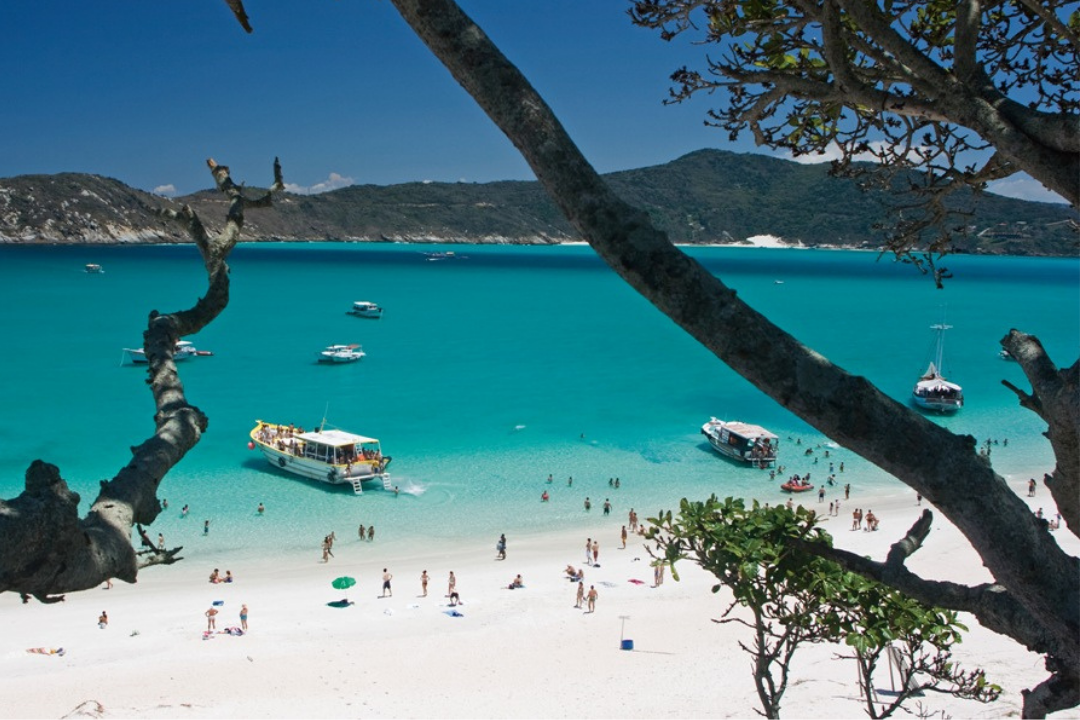 Praia do Farol, localizada na Ilha do Farol, em Arraial do Cabo. É possível ver pessoas na faixa de areia e embarcações no mar, além de um tronco de árvore em parte da foto.